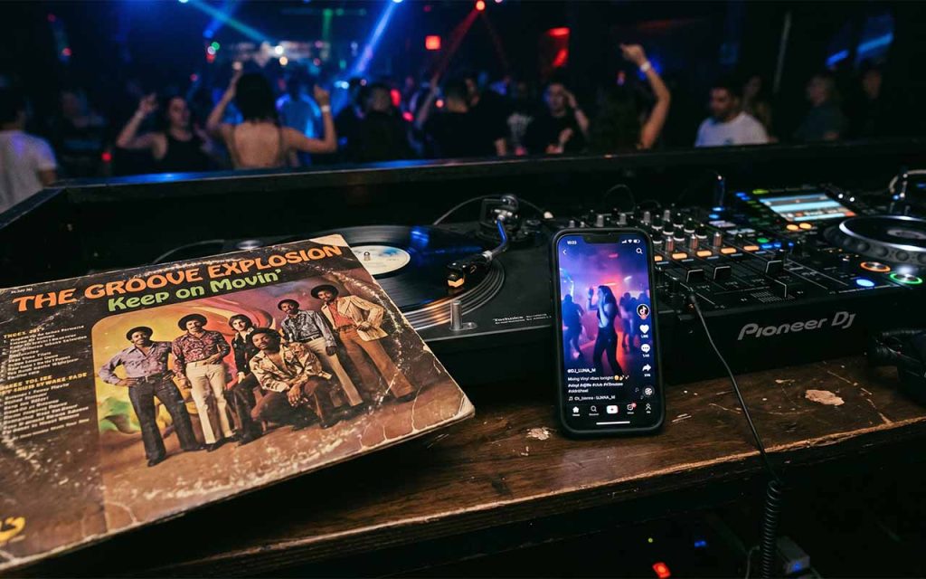 Ein abgenutztes Vinyl-Plattencover aus den 1970er Jahren liegt auf einem modernen DJ-Pult, direkt daneben leuchtet ein Smartphone mit der geöffneten TikTok-App in einem abgedunkelten Raum