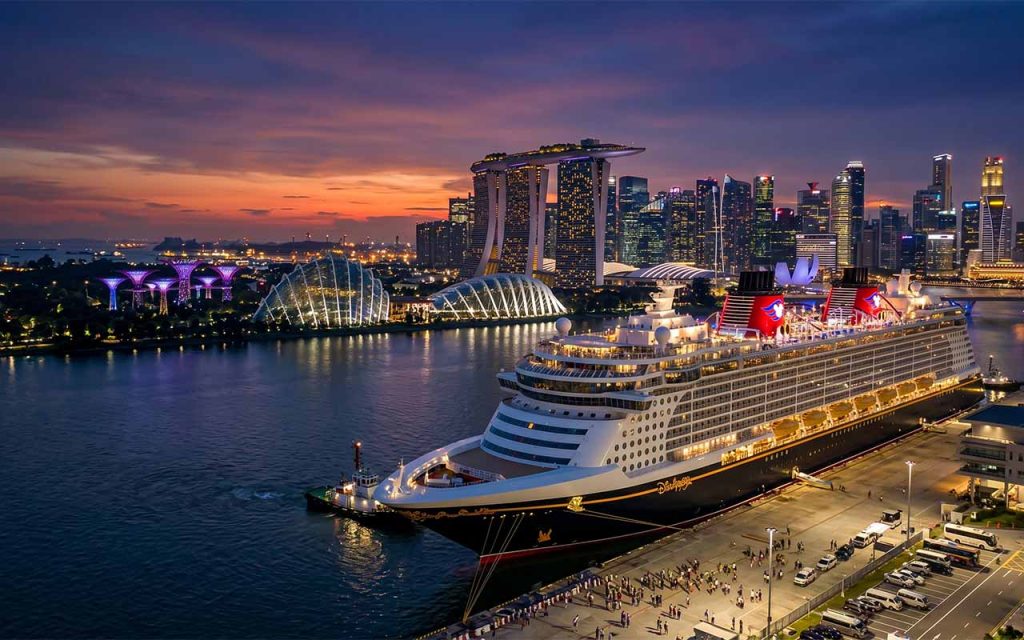 Eine majestätische Ansicht der Disney Adventure, die im glitzernden Hafen von Singapur anlegt, umgeben von der futuristischen Skyline von Marina Bay bei einsetzender Abenddämmerung